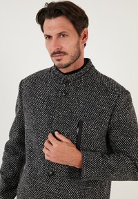 Abrigo de mezcla de lana en un patrón de espiga en blanco y negro. Cuenta con un cuello alto, dos botones frontales y un bolsillo en el pecho con cremallera.