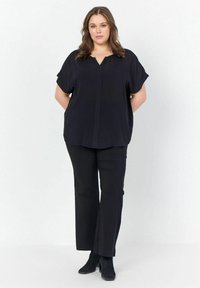 Blusa nera a maniche corte con scollo a V e dettagli di cucitura frontale sottili, abbinata a pantaloni neri e stivali alla caviglia, creando un completo coordinato.