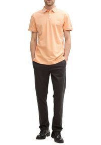 Chemise polo pêche avec un col, ornée d'un petit logo sur la poitrine. Associée à un pantalon gris foncé et des chaussures noires.