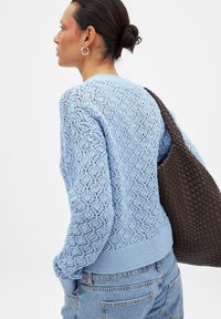 Svetlo modra pletena jopica z mustrom v obliki diamanta in rebrastim robom, v kombinaciji z temno rjavo tkano torbo.