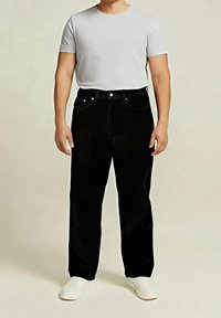 Homme portant un t-shirt blanc uni, un jean noir et des baskets blanches, debout devant un fond neutre.