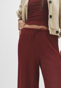 Pantaloni borgogna, strutturati, con una vestibilità rilassata, dotati di cintura regolabile. Abbinati a un top aderente e a una giacca beige con bottoni neri.