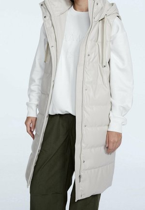 Personne portant un long gilet matelassé beige clair avec capuche, par-dessus un sweat-shirt blanc et un pantalon vert foncé, debout devant un fond uni.