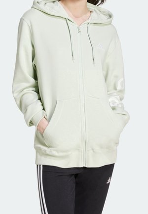 Person iført en lysegrøn hættetrøje med lynlås og kængurulommer samt sorte Adidas bukser med hvide striber.