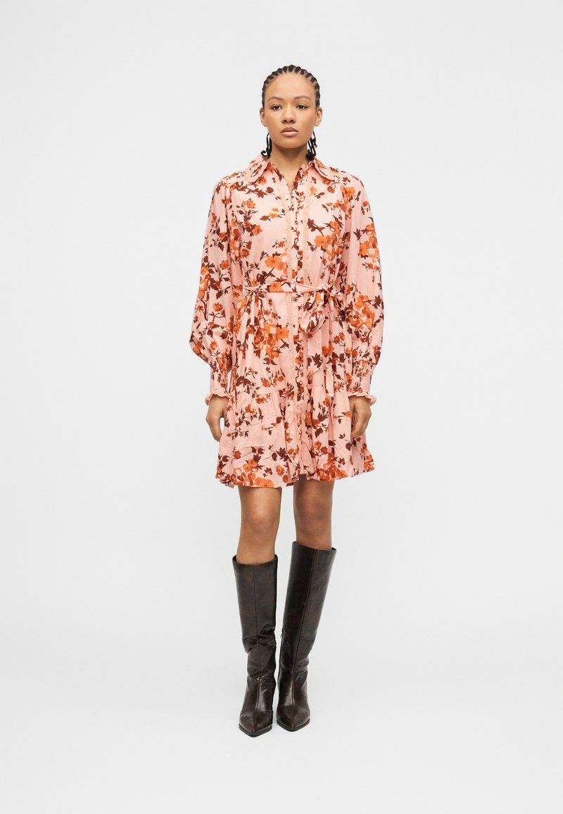 Γυναίκα με πλεξούδες, που φοράει μακρυμάνικο φόρεμα με ροζ λουλούδια και μαύρες μπότες έως το γόνατο, στέκεται μπροστά σε απλό φωτεινό φόντο.