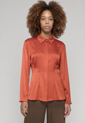 Femme aux cheveux bouclés portant un chemisier cintré à manches longues couleur rouille et un pantalon marron, debout devant un fond clair uni.