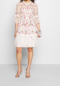 Femme portant une robe blanche longueur genou avec broderies florales roses, manches transparentes, tenant une pochette argentée, et sandales à talons hauts argentées.
