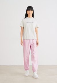 Svetlošedé bavlnené tričko s nápisom "Calvin Klein Jeans". Mäkké ružové, voľne strihané teplákové nohavice s elastickými manžetami. Biele tenisky dopĺňajú celý outfit.