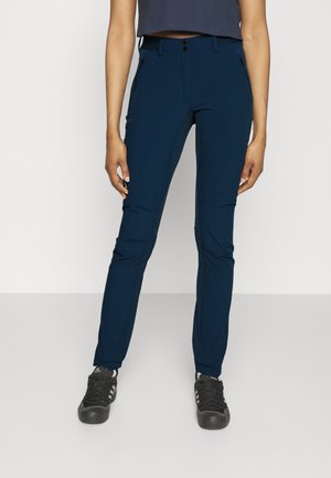 Person iført marineblå slim-fit bukser med lynlåslommer og sorte Adidas sneakers, stående mod en ensfarvet baggrund.