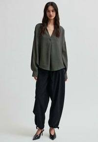 Blusa verde oliva a maniche lunghe con scollatura a V e dettagli con bottoni, abbinata a pantaloni neri larghi che si stringono alle caviglie. Inclusi tacchi neri.