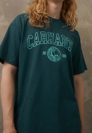 Person med langt krøllet hår iført en mørkegrøn Carhartt T-shirt med lyseblå tekst og logo mod en grå baggrund.