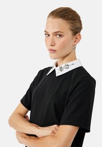 Camisa negra de manga corta con un cuello blanco adornado con embellishments de cristal decorativos, que presenta una silueta ajustada y una textura suave.