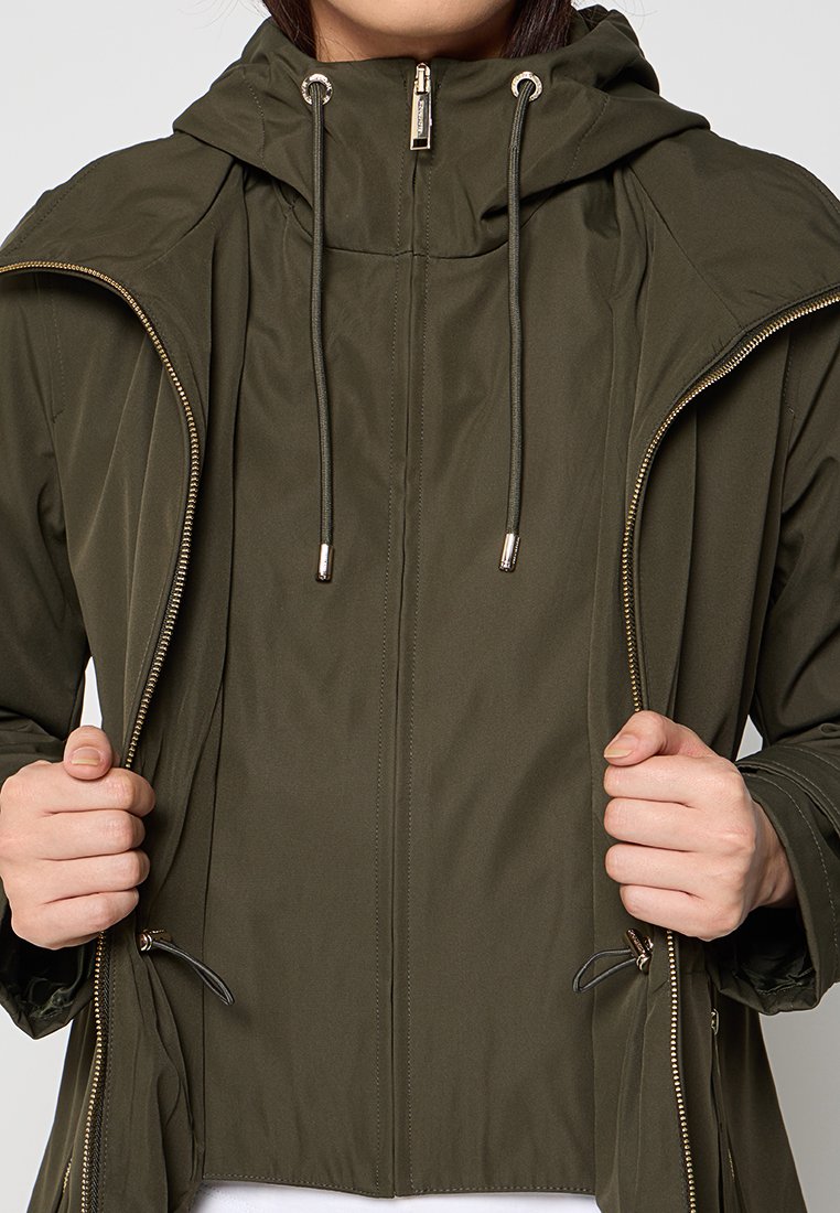 Kobieta w oliwkowej kurtce z kapturem, zapinanej na zamek, trzymająca brzegi kurtki częściowo rozpiętej w okolicy klatki piersiowej.