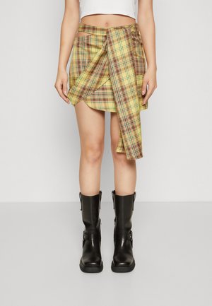 Plaid wrap skirt in yellow with brown accents, featuring a high waist and asymmetric hem. Paired with black knee-high boots.