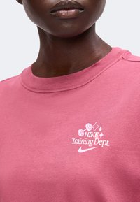 Крупен план на човек, носещ розова риза с бял текст "Nike Training Dept." и графика на щанга на гърдите.