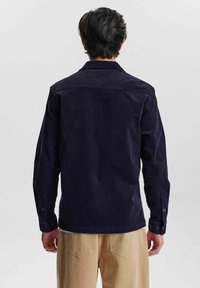 Cămașă din velur navy cu mâneci lungi, manșete cu nasturi și guler ajustat, purtată cu pantaloni de culoare deschisă. Material texturat, design simplu.