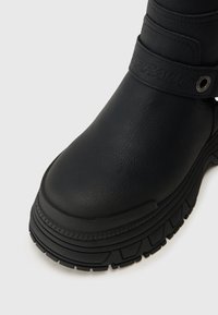 Czarny skórzany but z zaokrąglonym noskiem, grubą teksturowaną podeszwą i dekoracyjnym paskiem wytłoczonym napisem "BUFFALO" w pobliżu kostki.