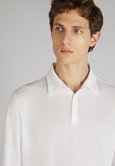 Witte polo met een structuurstof, voorzien van een klassieke kraag en een drieknoopsluiting. Model toont het shirt van borsthoogte omhoog.