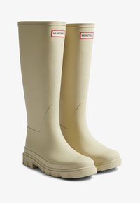 Bottes montantes en caoutchouc beige avec une semelle texturée, un design courbé à la cheville et un logo "HUNTER" rouge et blanc sur le côté supérieur.