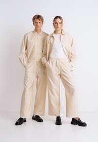 Σετ μπεζ με ρίγες που περιλαμβάνει πουκάμισο με μακριά μανίκια και κουμπιά, καθώς και παντελόνια με χαλαρή εφαρμογή, και τα δύο με ελαστικές ζώνες μέσης και μινιμαλιστικό σχεδιασμό.