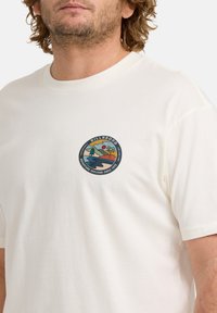 Cremefarbendes Baumwoll-T-Shirt mit einem runden, mehrfarbigen Grafiklable auf der linken Brust. Kurze Ärmel und ein klassischer Rundhalsausschnitt.