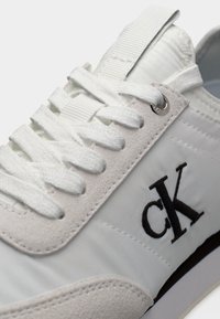 Λευκό αθλητικό παπούτσι με λευκά κορδόνια, σουέντ λεπτομέρειες και μαύρο λογότυπο "ck" στην πλευρά, εστιασμένο στο πάνω μπροστινό μέρος.