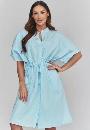 Kobieta o długich ciemnych włosach, ubrana w jasnoniebieską, krótką sukienkę do kolan z taliowaną talią i stojącym kołnierzykiem.