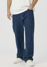 Modre kavbojke iz jeans tkanine z rahlo sproščenim krojem, vidnimi šivi in sprednjimi žepi. Oblečene z belimi supergami in enostavno belo majico.