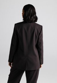 Black tailored blazer with structured shoulders, a single back vent, and buttoned cuffs. Smooth fabric with a sleek design.