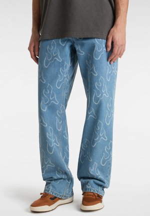 Blå denimjeans med lette flammemønstre båret med brune sneakers og en mørkegrå t-shirt på en stående person.