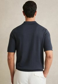 Tamsiai mėlynas trumpomis rankovėmis polo marškinėliai su minkšta tekstūra, klasikinis apykaklė ir mėgtukai, derinami su beige kelnėmis ir minimalistiniu dizainu.
