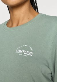 Grønn bomulls-T-skjorte med rund hals og hvit trykt logo som sier "LIMITLESS" med "ONLY PLAY ENERGY" over.