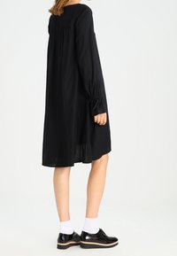 Robe noire fluide avec des manches longues et des détails froncés aux épaules, associée à des chaussures en cuir verni noires et des chaussettes blanches à la cheville.