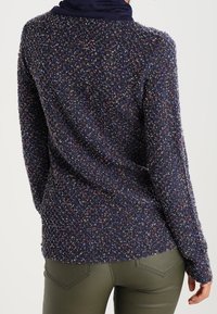 Mörkblå texturerad tröja med flerfärgade fläckar, rund halsringning och långa ärmar, i kombination med gröna åtsittande byxor.