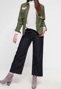 Oliivinvihreä työkalutakki, jossa on kangasmerkkejä, kermaan vivahtava neule, tummansiniset leveälahkeiset farkut, joissa on viimeistelemätön helma, ja harmaat nilkkurit.