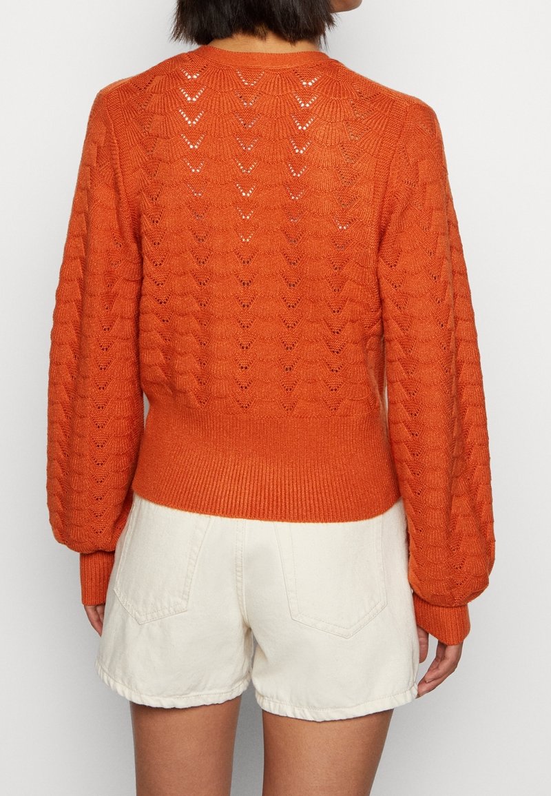 Femme portant un pull en tricot orange avec un motif festonné et un short blanc taille haute, vue de dos.