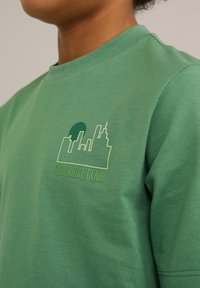 Žalias medvilninis marškinėlis su grafiniu dizainu, vaizduojančiu miesto panoramą ir šviesiai žalią ratą. Taip pat spausdintas tekstas "BLUE RIDGE DENIM".