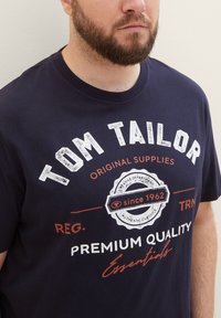 Granatowa bawełniana koszulka z białym i pomarańczowym napisem, z logo "TOM TAILOR", wzorem pieczęci i napisem "PREMIUM QUALITY". Krótkie rękawy.