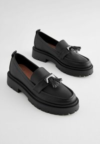 Black leather loafers with textured finish, chunky rubber sole, and tassel detail on strap. Rounded toe and pull tab at heel.
