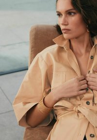 Cămașă bej cu mânecă scurtă și guler, cu detalii cu nasturi și un talie legată. Modelul poartă o brățară țesută. Fundal din catifea.