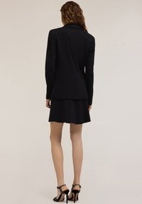 Blazer nero su misura e gonna a pieghe, entrambi realizzati in tessuto liscio. Sandali con tacco alto e cinturini alla caviglia. Linee pulite e dettagli minimi.