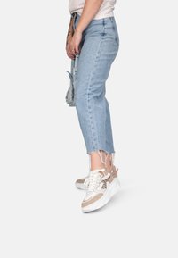 Jeans de denim azul claro con dobladillo deshilachado, combinados con zapatillas blancas y beige con un diseño texturizado. Un bolso en la mano completa el conjunto.