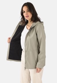 Chaqueta con capucha beige ligera, forro interior negro, cintura elástica y bolsillos con cremallera. Textura suave y diseño moderno.