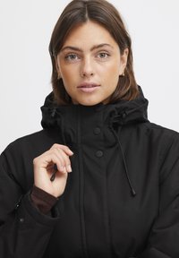 Veste noire imperméable avec un col montant, des boutons à l'avant et une capuche avec cordon, présentant une texture lisse et des détails de conception pratiques.