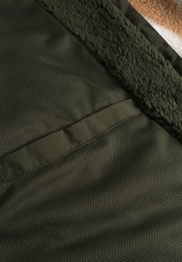 Chaqueta verde oliva con un cuello de forro polar texturizado y una tela de malla ligera. Presenta un bolsillo en el pecho horizontal y forro suave.