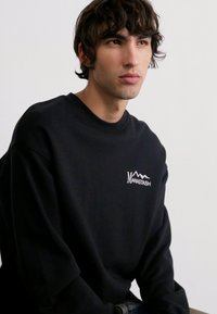 Black crewneck sweatshirt made of cotton, featuring a small white "Manastash" logo and mountain graphic on the left chest.