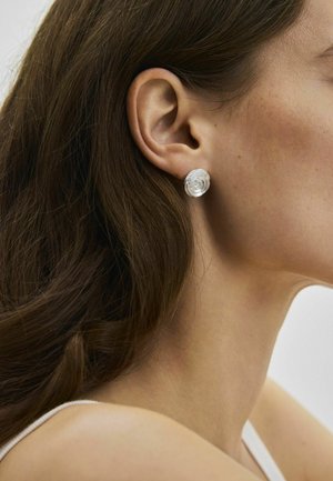 Femeie cu păr lung și șaten, purtând o cercei spiral translucizi, prezentată în profil, concentrându-se pe ureche și linia maxilarului.