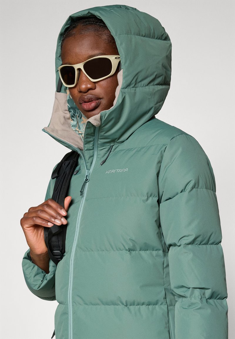 Grüne Daunenjacke mit Kapuze, ausgestattet mit einem Frontreißverschluss und eingeprägtem Logo. Enthält ein leichtes Innenfutter und wird über einem schwarzen Rucksack getragen.