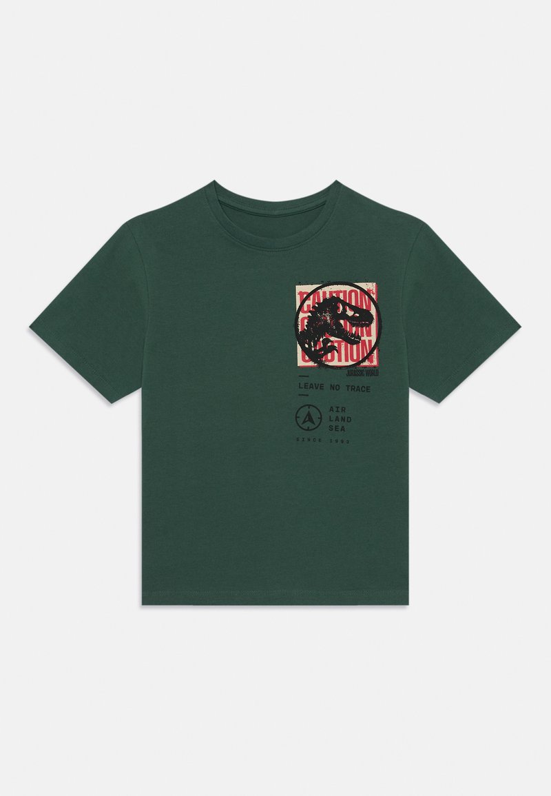 Camiseta de algodón verde con un gráfico de dinosaurio, fondo rojo de advertencia y el texto "No Dejes Rastro" con un logo debajo. Mangas cortas.