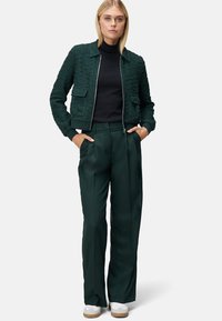 Bomber vert texturé avec fermeture éclair à l'avant, associé à un pantalon sombre vert sur mesure et des baskets blanches. Deux poches avant visibles.